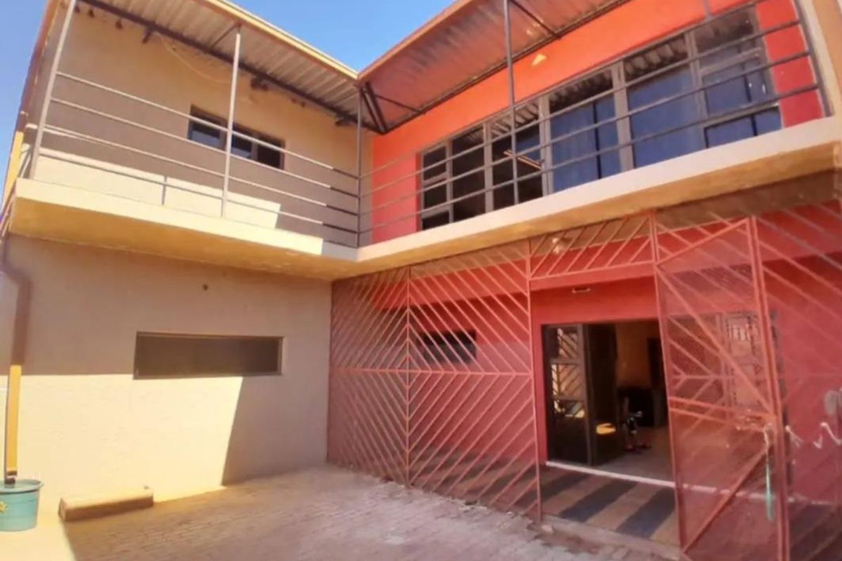 Rear exterior view of a two-story house featuring a paved courtyard, upper-level balcony, and metal security gate entrance.