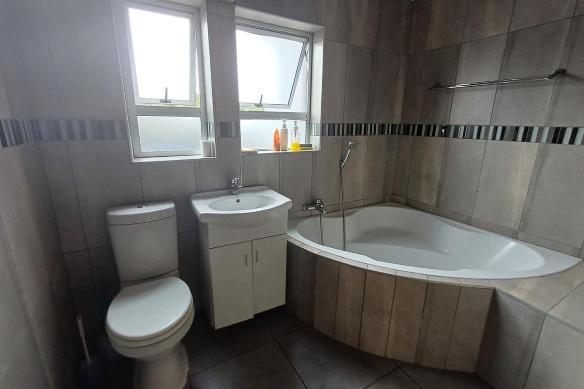 A functional bathroom featuring a corner bathtub, vanity basin, toilet, and neutral wall and floor tiling with natural light from casement windows.
