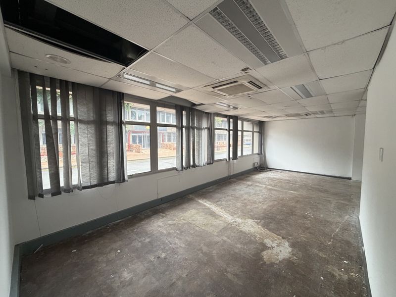 An empty industrial office space featuring large windows, suspended ceilings with integrated lighting, and ducted air conditioning, requiring refurbishment of flooring and ceiling tiles.