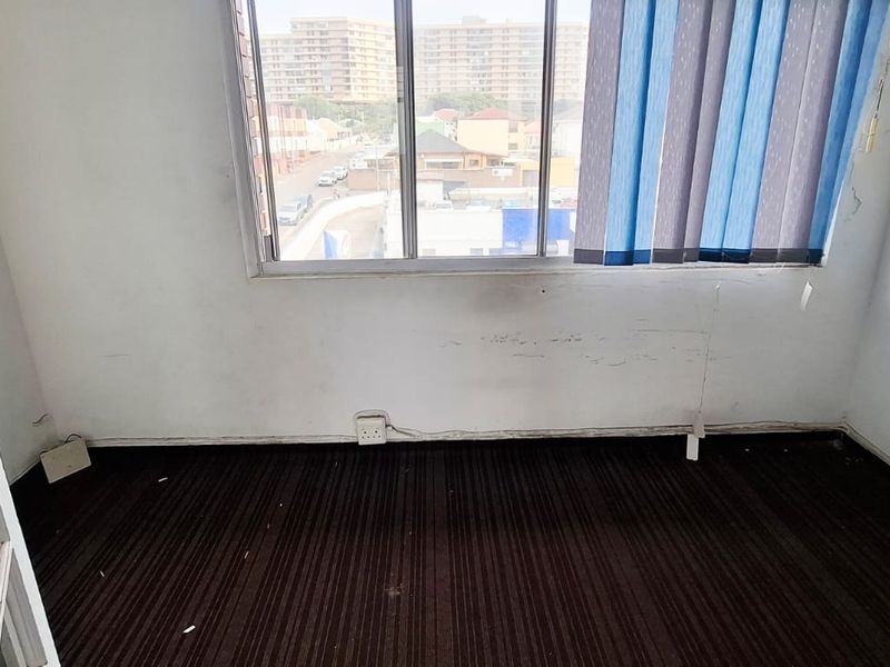 An empty office space featuring large windows with vertical blinds, dark carpet flooring, and visible wall wear requiring maintenance.