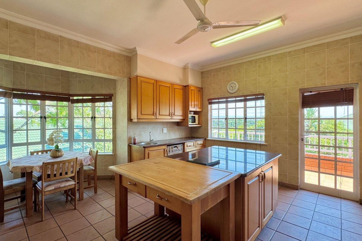 Spacious kitchen featuring wooden cabinetry, a central island with granite countertops, tiled flooring, and large windows providing ample natural light and garden views.