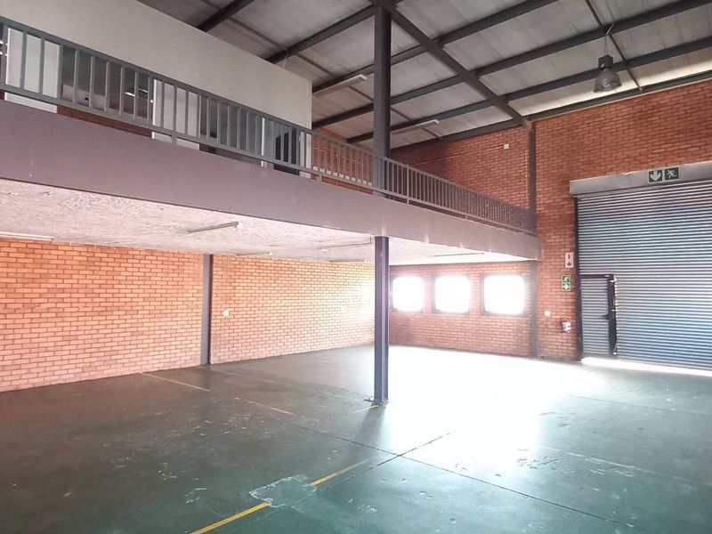 Spacious industrial warehouse featuring high ceilings, exposed brick walls, a mezzanine level with metal railing, and a large roller shutter door for easy access.