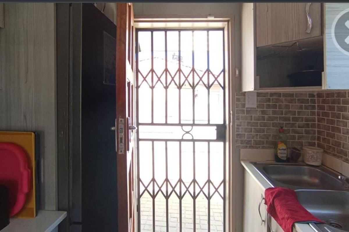 A functional kitchen area featuring a double stainless steel sink, tiled backsplash, and built-in cabinetry, with a security gate leading to the exterior.