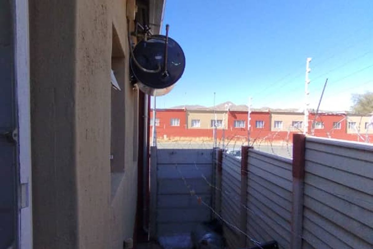 A narrow rear exterior side passage featuring a wall-mounted electric water heater, paved flooring, and perimeter security fencing.