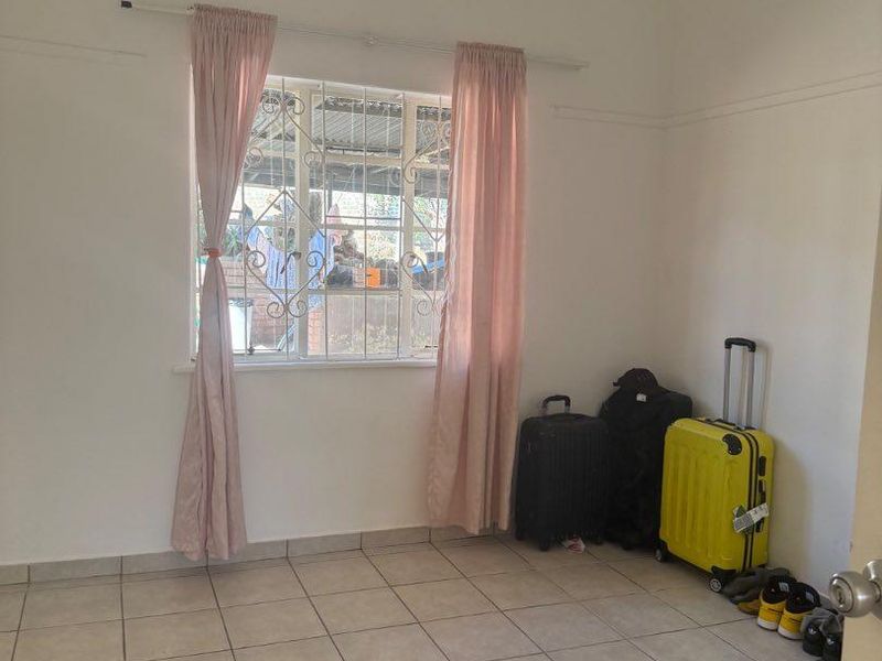 A simple, vacant bedroom featuring neutral tiled flooring, a window with security bars, and a curtain rod, offering a clean canvas for personalization.