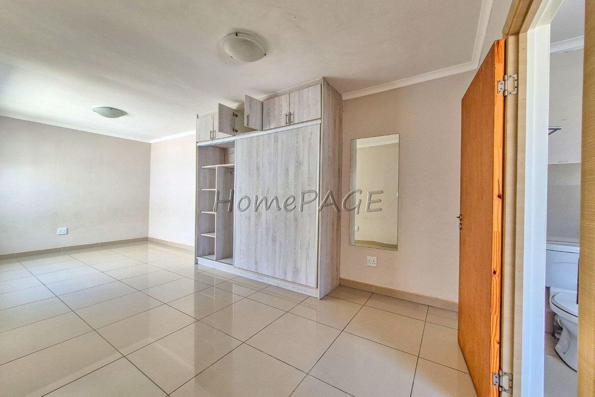 A bright, empty bedroom featuring neutral tiled flooring, built-in wooden cabinetry, and a wall-mounted mirror, with an adjacent bathroom entrance.
