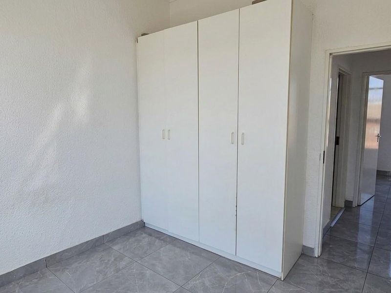 A clean, empty bedroom featuring modern grey tiled flooring and a white built-in wardrobe, with an adjacent hallway visible through the doorway.