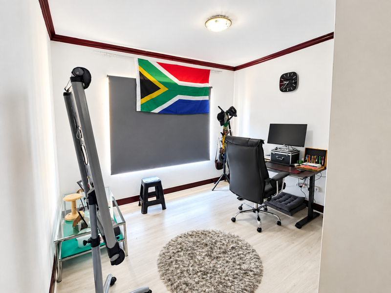 A functional home office space featuring light-toned laminate flooring, dark decorative ceiling cornicing, and a large window with a fitted roller blind.