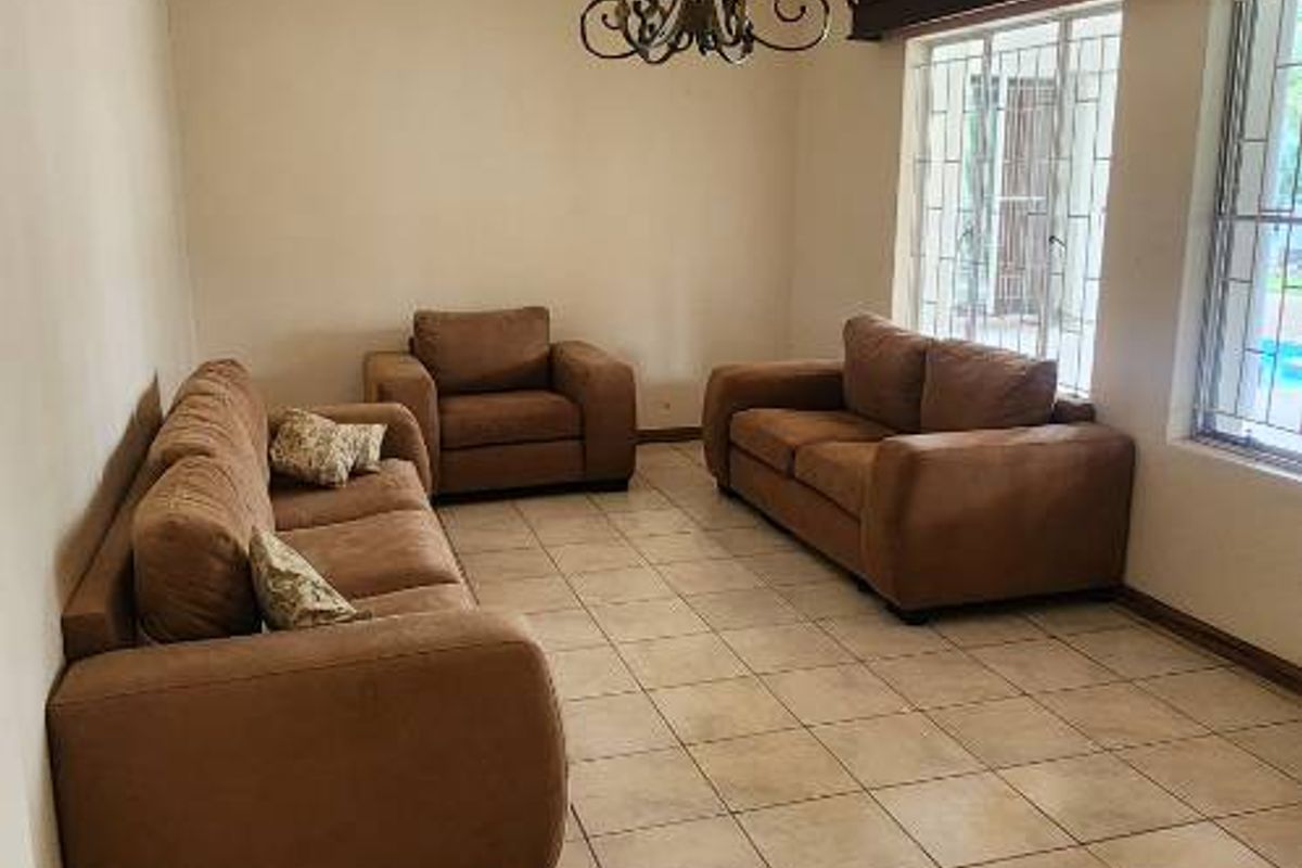 A spacious living room featuring neutral tiled flooring, large windows with security bars, and a decorative chandelier.