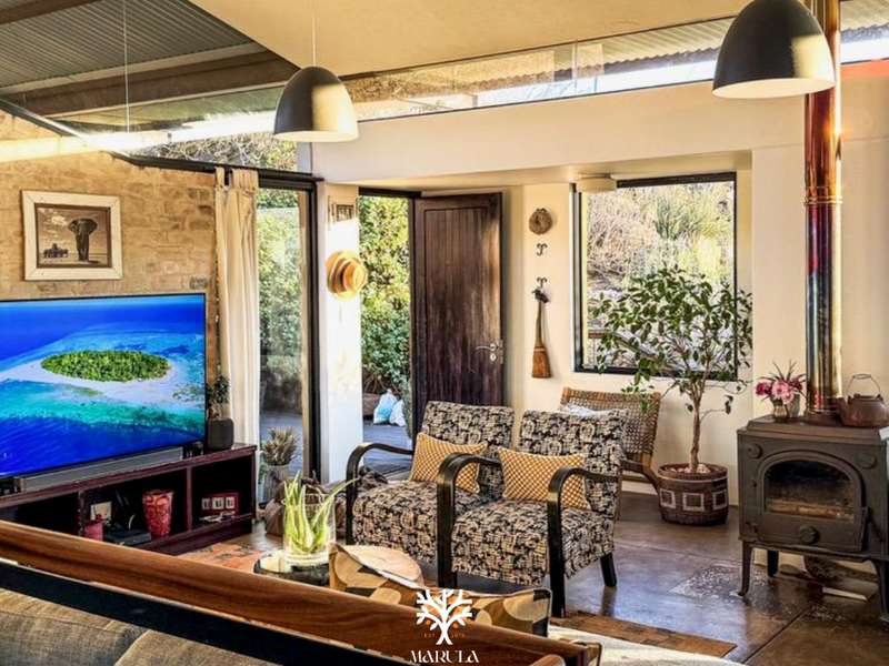 A cozy living room featuring an exposed stone wall, a wood-burning stove, concrete floors, and large windows that provide natural light and garden views.