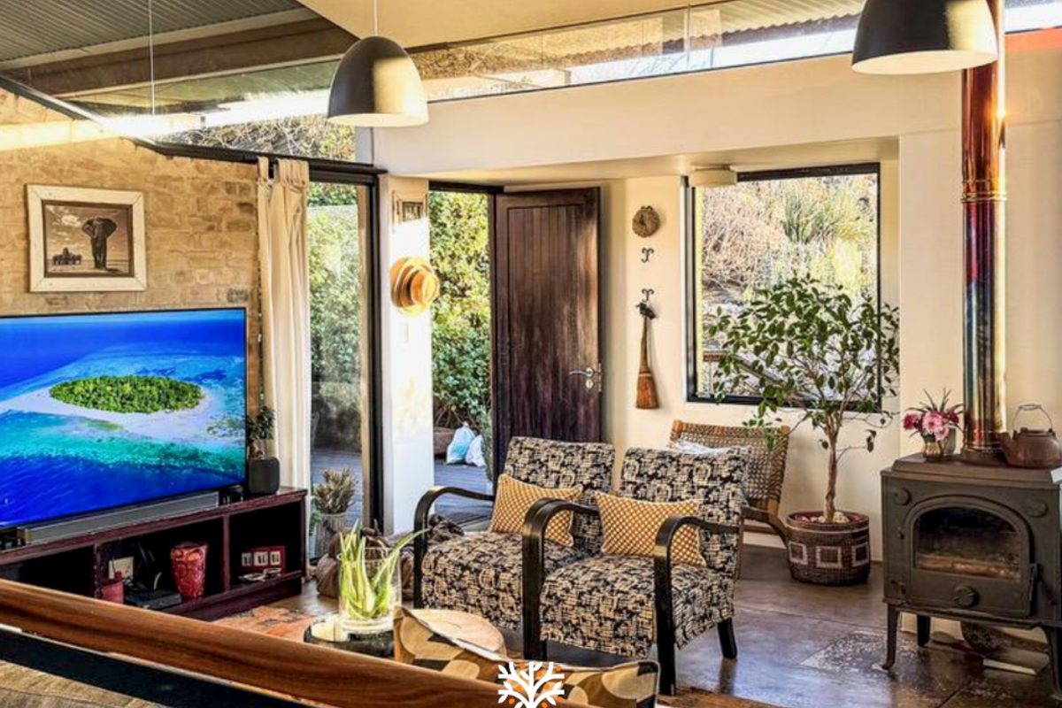 A cozy living room featuring an exposed stone wall, a wood-burning stove, concrete floors, and large windows that provide natural light and garden views.