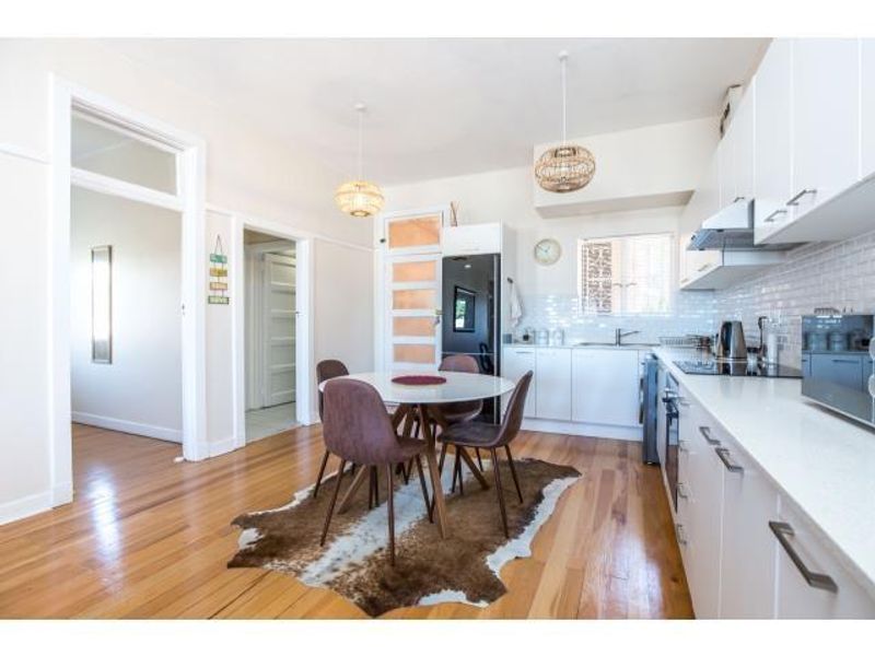 A bright, modern kitchen featuring white cabinetry, stone countertops, subway tile backsplash, and polished hardwood flooring, seamlessly integrated with a dining area.