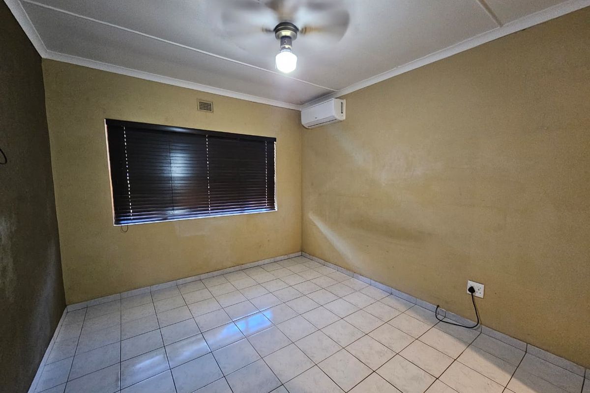 An empty bedroom featuring tiled flooring, a ceiling fan, a wall-mounted air conditioning unit, and dark window blinds.