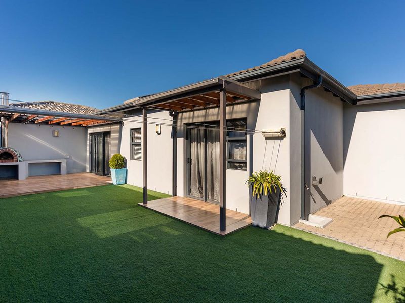 Rear exterior view featuring a covered patio with timber decking, an integrated pizza oven, and a well-maintained artificial lawn area.