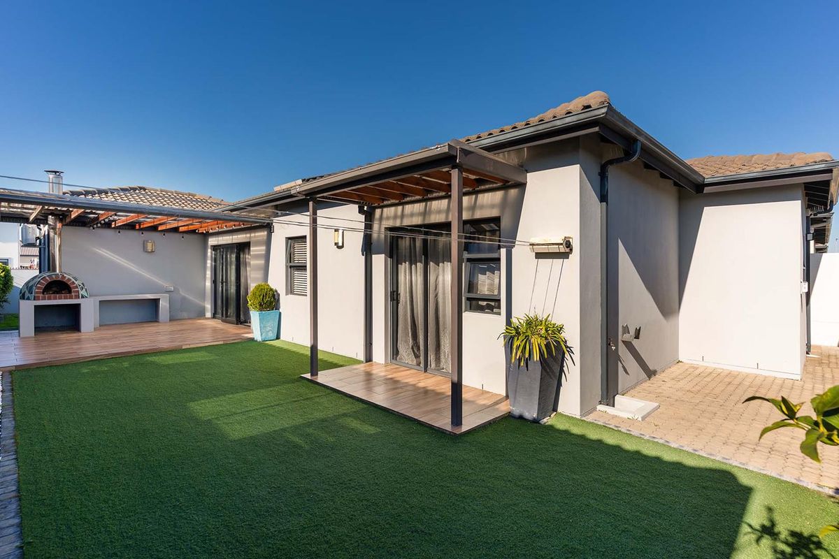Rear exterior view featuring a covered patio with timber decking, an integrated pizza oven, and a well-maintained artificial lawn area.