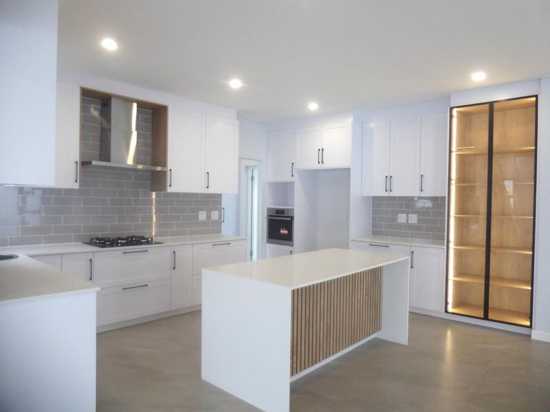 Modern, newly renovated kitchen featuring white cabinetry, a central island with timber detailing, subway tile backsplash, built-in appliances, and integrated display shelving.