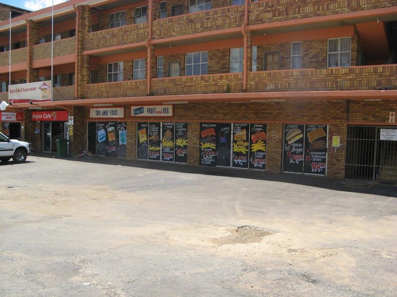 Exterior view of a multi-story apartment building featuring ground-floor commercial retail spaces and an asphalt parking area.