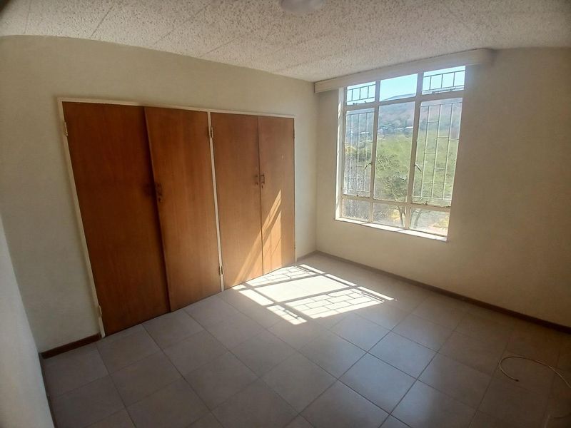 A bright, empty bedroom featuring tiled flooring, built-in wooden cupboards, and a large window providing natural light and exterior views.