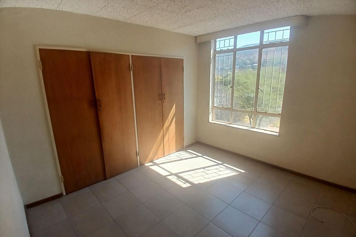 A bright, empty bedroom featuring tiled flooring, built-in wooden cupboards, and a large window providing natural light and exterior views.