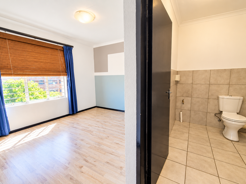 A bright, empty bedroom featuring laminate flooring and an adjacent tiled bathroom with a standard toilet fixture.