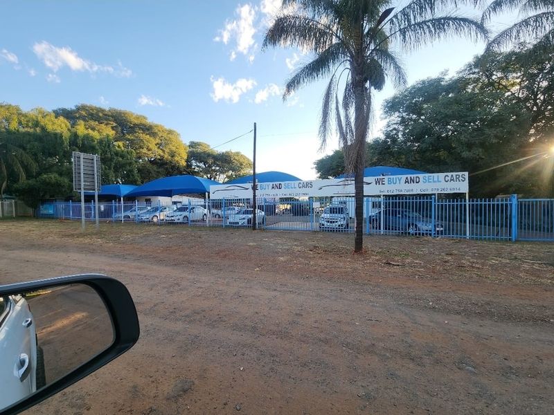 Front view of a car dealership property featuring blue perimeter fencing, shade cloth carports, and a paved vehicle display area.