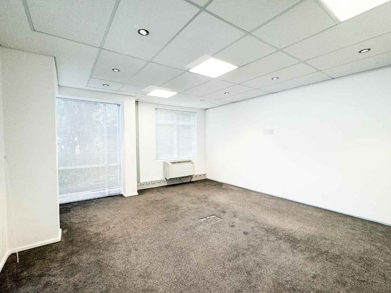 A clean, vacant office space featuring a suspended ceiling with recessed lighting, carpeted flooring, and a wall-mounted air conditioning unit.