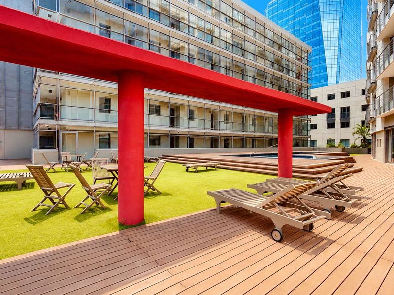 A well-maintained apartment complex pool area featuring timber decking, artificial grass, and a striking red structural pergola.