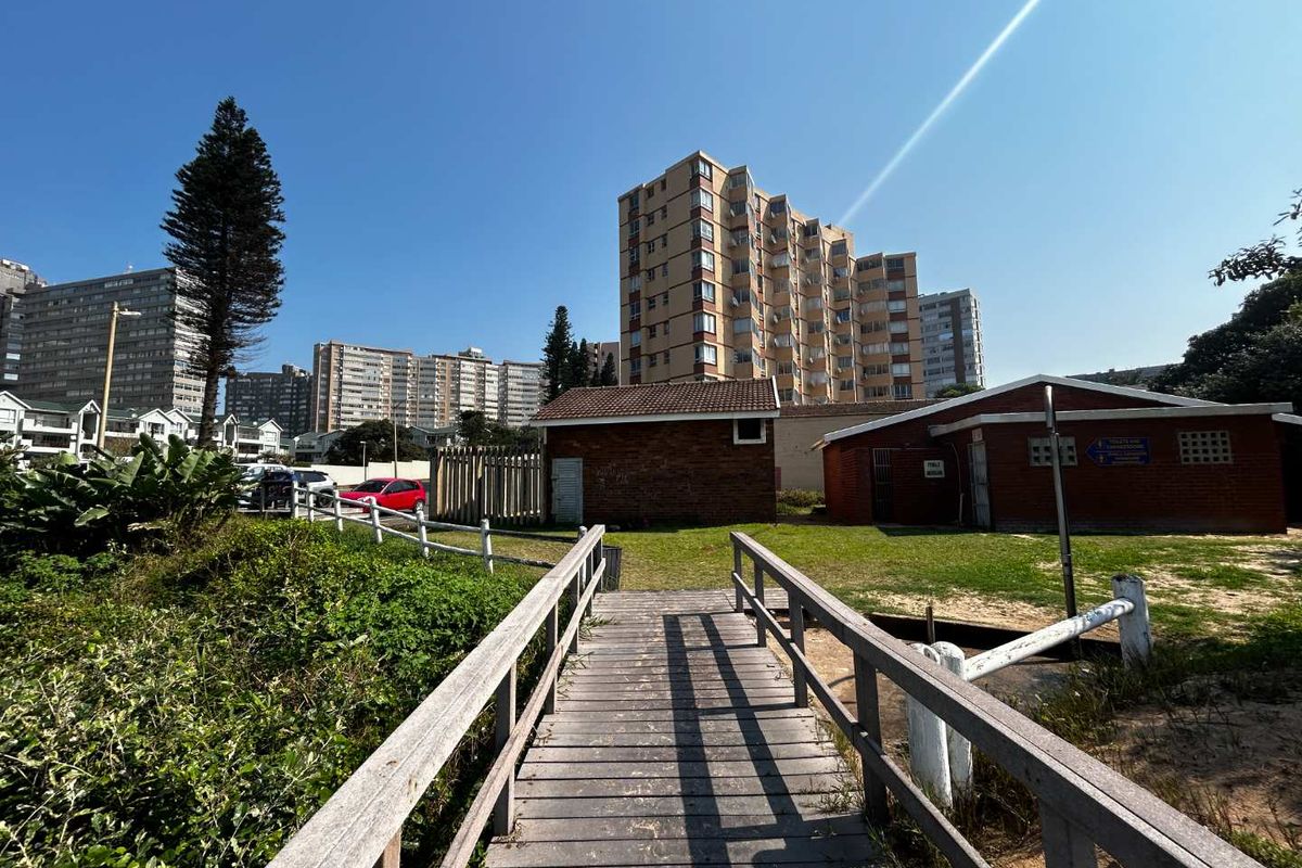 A wooden boardwalk path leading towards a communal area with brick outbuildings and surrounding apartment blocks in a coastal setting.
