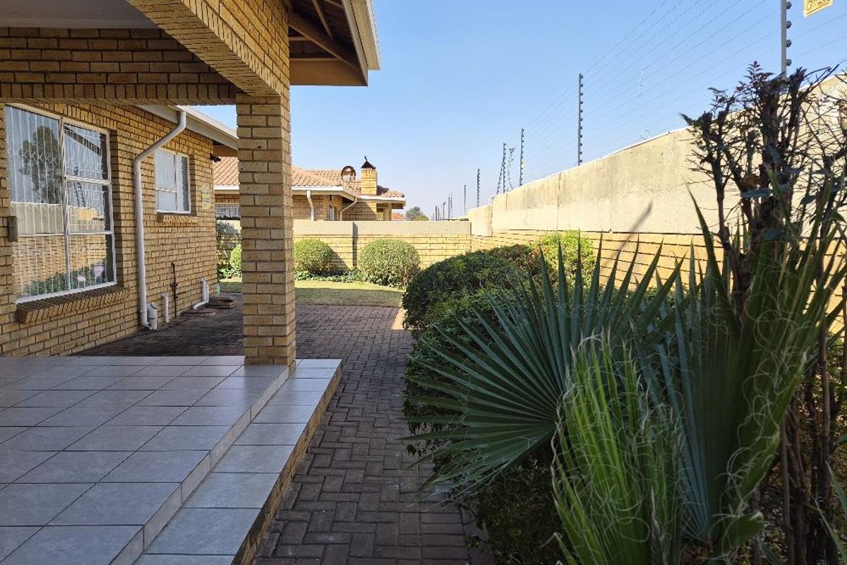 A view of the rear exterior featuring a covered tiled patio, brick facade, paved walkway, and secure boundary wall with electric fencing.