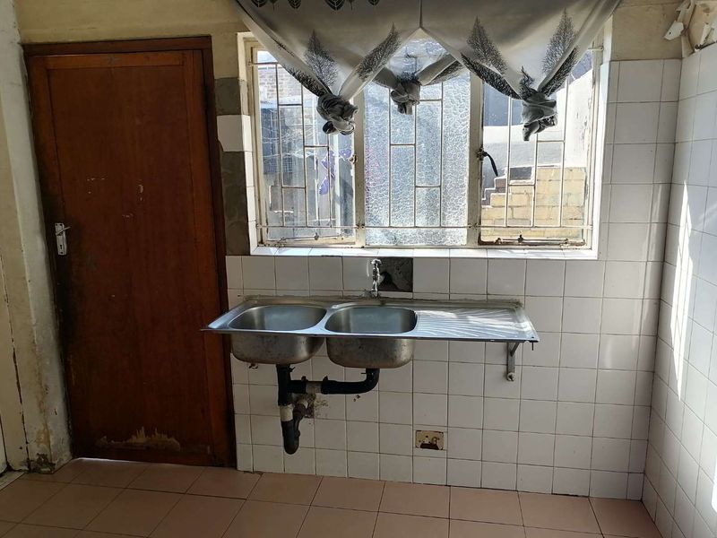 A basic kitchen area featuring a double stainless steel sink, tiled splashback, and security-barred window, requiring significant cosmetic updates and maintenance.