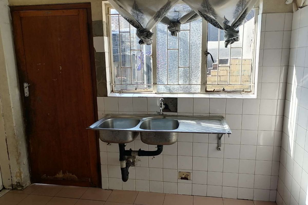 A basic kitchen area featuring a double stainless steel sink, tiled splashback, and security-barred window, requiring significant cosmetic updates and maintenance.
