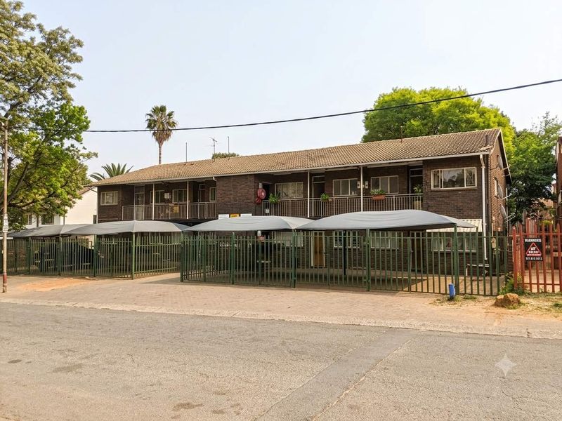 Front exterior view of a multi-story brick apartment building featuring covered parking bays and secure perimeter fencing.