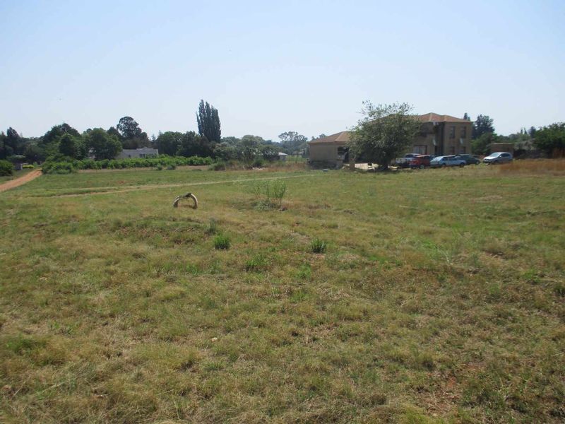 A wide-angle view of a vacant residential plot featuring open grassland with surrounding trees and neighboring properties in the distance.
