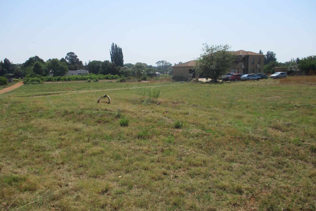 A wide-angle view of a vacant residential plot featuring open grassland with surrounding trees and neighboring properties in the distance.