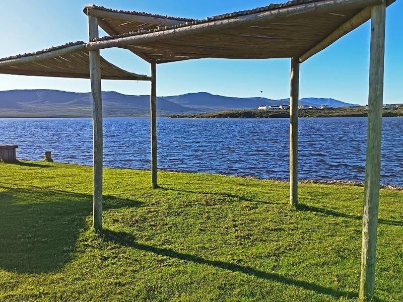 A rustic timber pergola structure situated on a grassy lawn overlooking a large lake with mountain views in the distance.