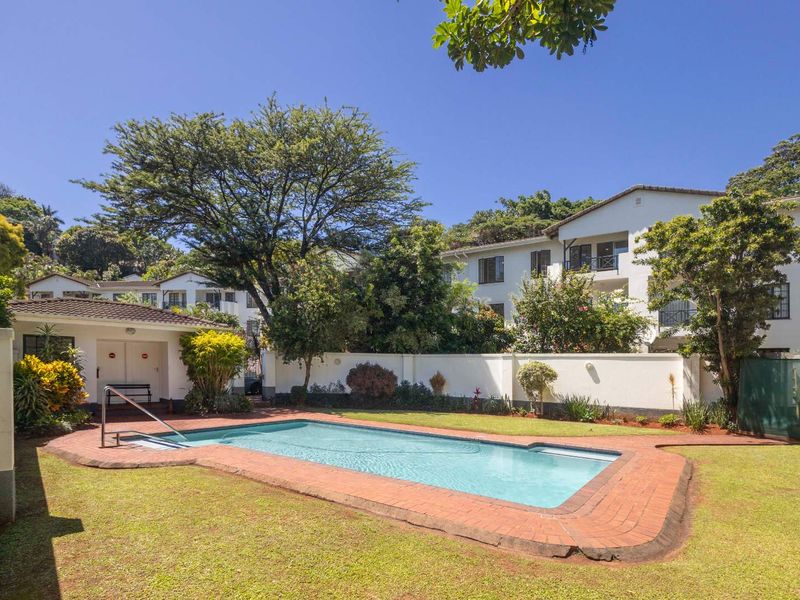 A well-maintained outdoor pool area featuring a rectangular swimming pool with brick coping, surrounded by a lush garden and perimeter wall.