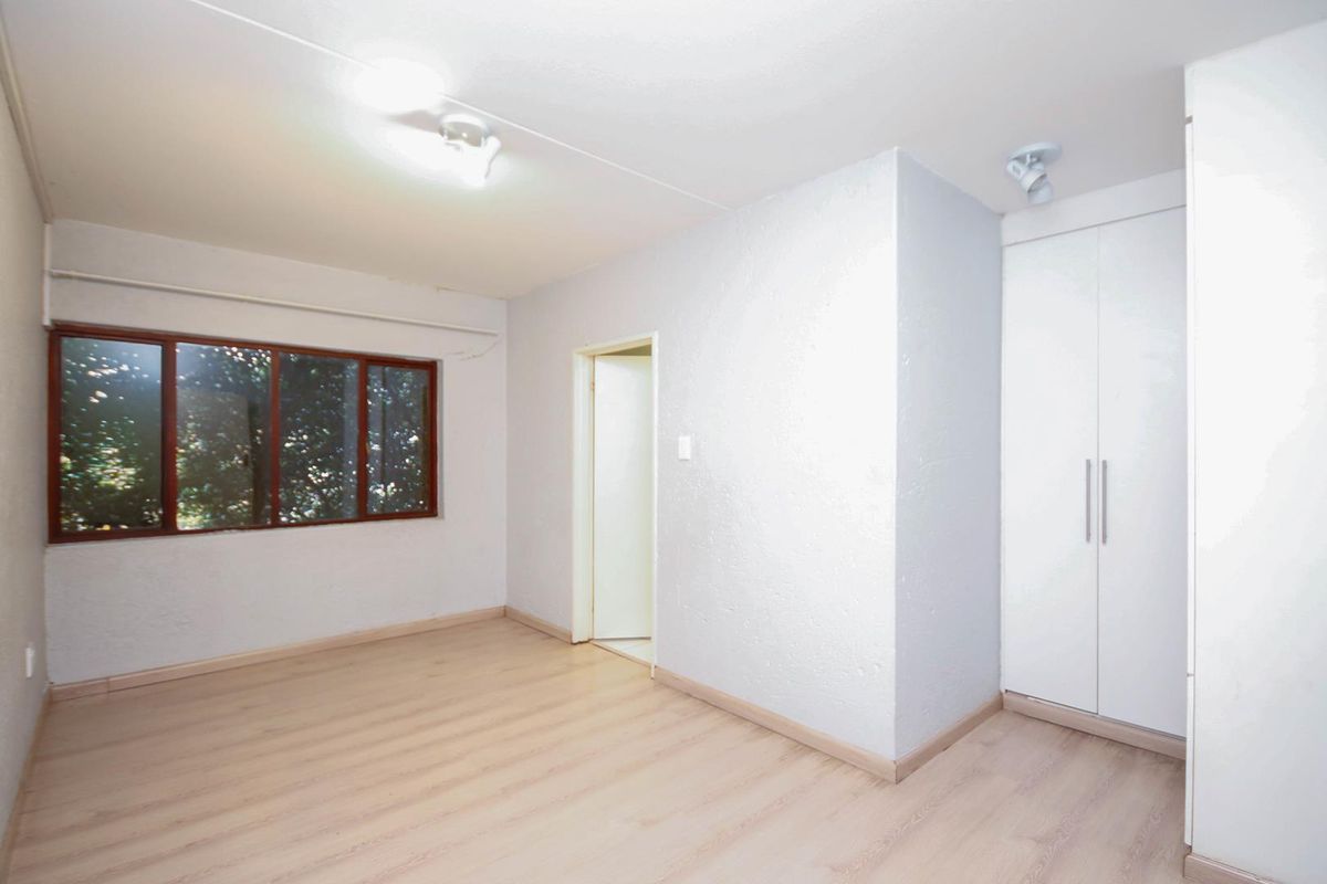 A bright, empty bedroom featuring light-toned laminate flooring, a built-in wardrobe, and a large window providing natural light.