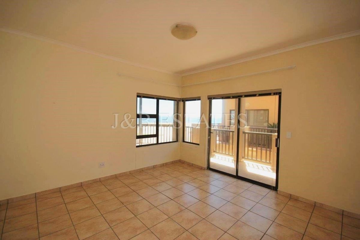An empty, well-lit living room featuring neutral tiled flooring, large aluminium-framed windows, and a sliding glass door providing access to an outdoor balcony area.