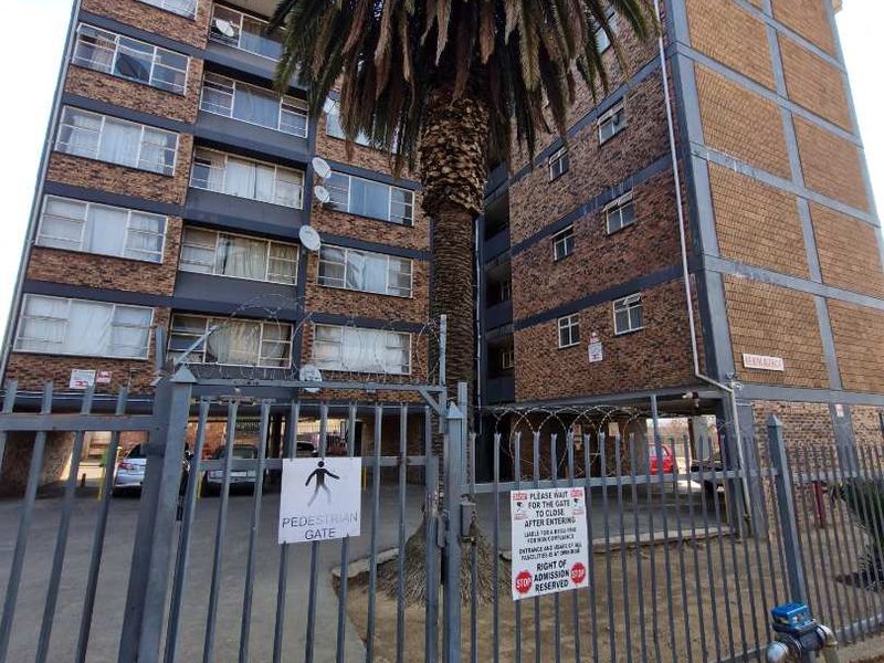 Exterior view of a multi-story apartment building featuring a brick facade, secure metal perimeter fencing, and a pedestrian access gate.