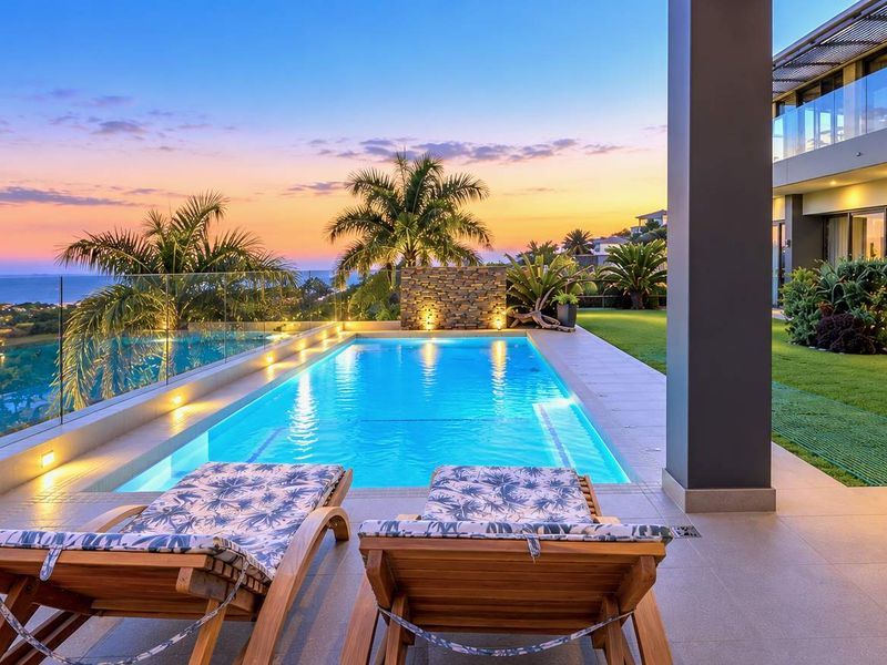 Luxury outdoor pool area featuring a modern swimming pool, glass balustrades, stone feature wall, and expansive views at sunset.