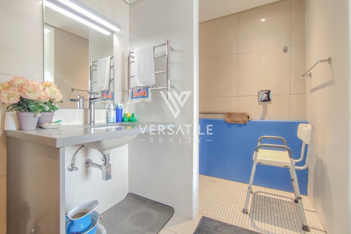 A functional bathroom featuring a vanity with integrated basin, wall-mounted mirror, heated towel rail, and a walk-in shower area with grab rails.