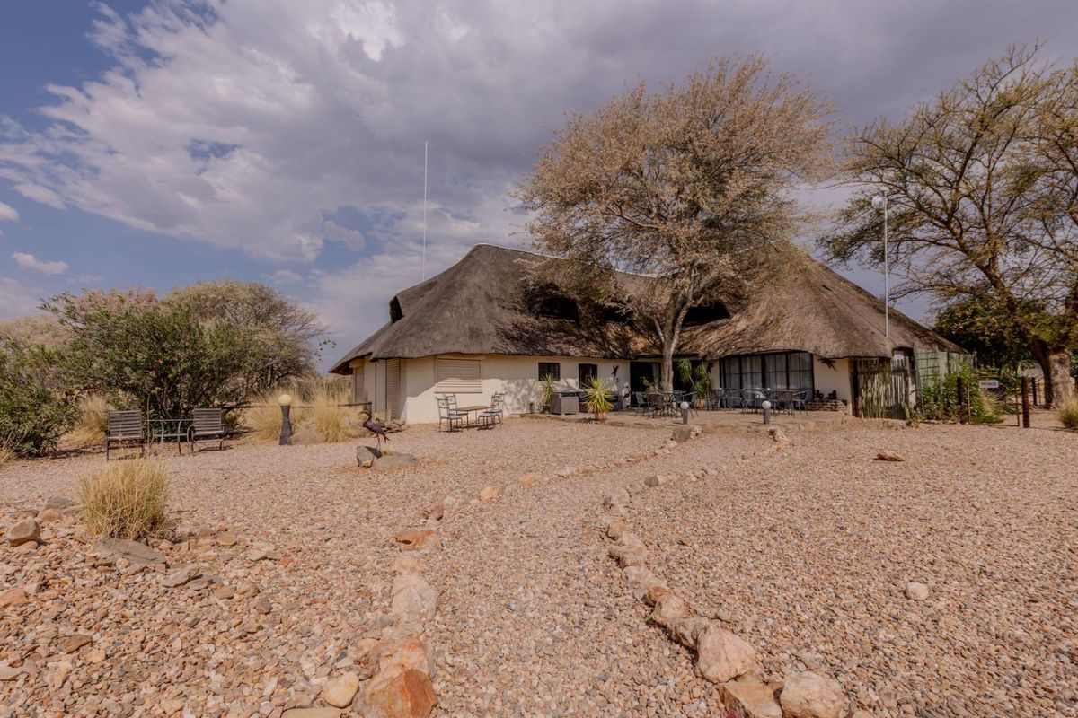 Rear view of a lodge featuring a traditional thatched roof, stone-paved patio area, and expansive gravel garden surroundings.