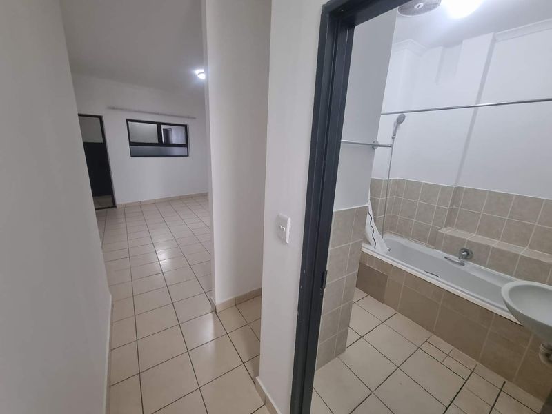 A view from a hallway looking into an empty bathroom with a tiled bathtub and sink, featuring neutral floor tiles throughout.