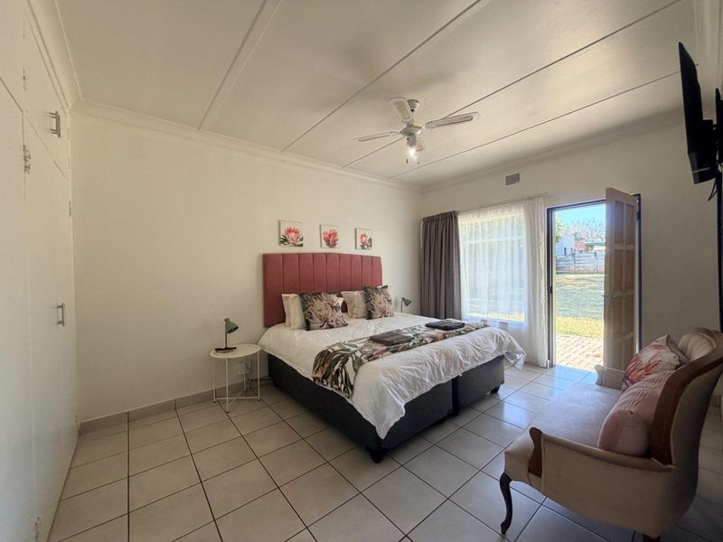 A bright bedroom featuring tiled flooring, built-in cupboards, a ceiling fan, and direct access to the garden through a sliding door.