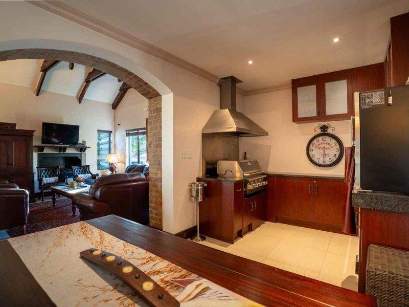 Open-plan living area featuring a kitchen with built-in braai, granite countertops, and wooden cabinetry, flowing into a cozy lounge with a fireplace and exposed beams.