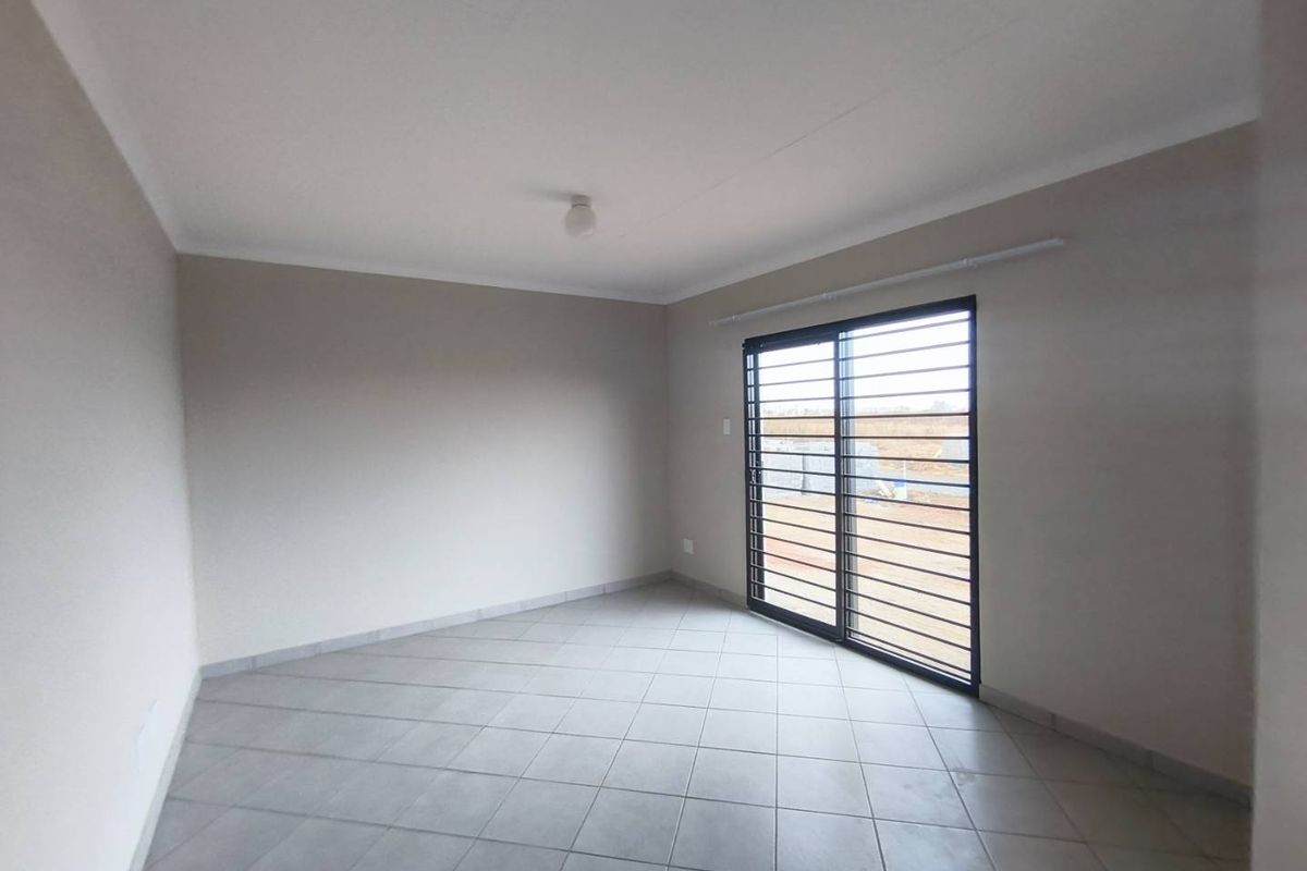 A bright, empty living room featuring neutral tiled flooring, plain walls, and a sliding glass door with security bars.