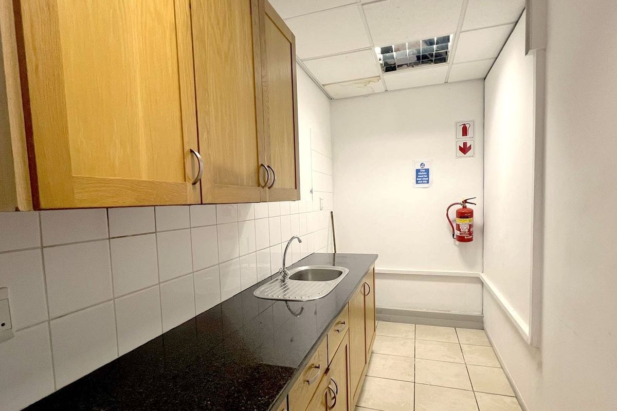 A functional office kitchenette featuring built-in wooden cabinetry, a dark laminate countertop with a stainless steel sink, and tiled flooring.