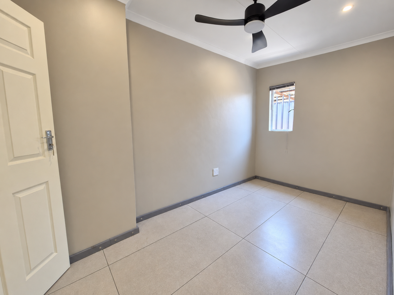 A clean, empty bedroom featuring neutral wall paint, large floor tiles, a ceiling fan, and a window with a blind.