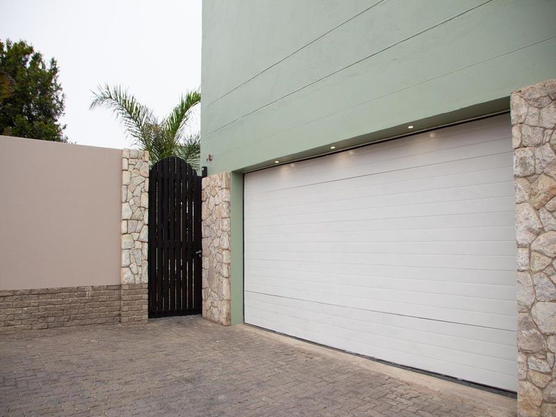 Modern front facade featuring a clean sectional garage door, stone-clad pillars, and a secure pedestrian gate with paved driveway access.