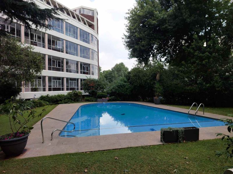 A well-maintained communal swimming pool area set within a landscaped garden, featuring paved surrounds and direct access to the apartment building.